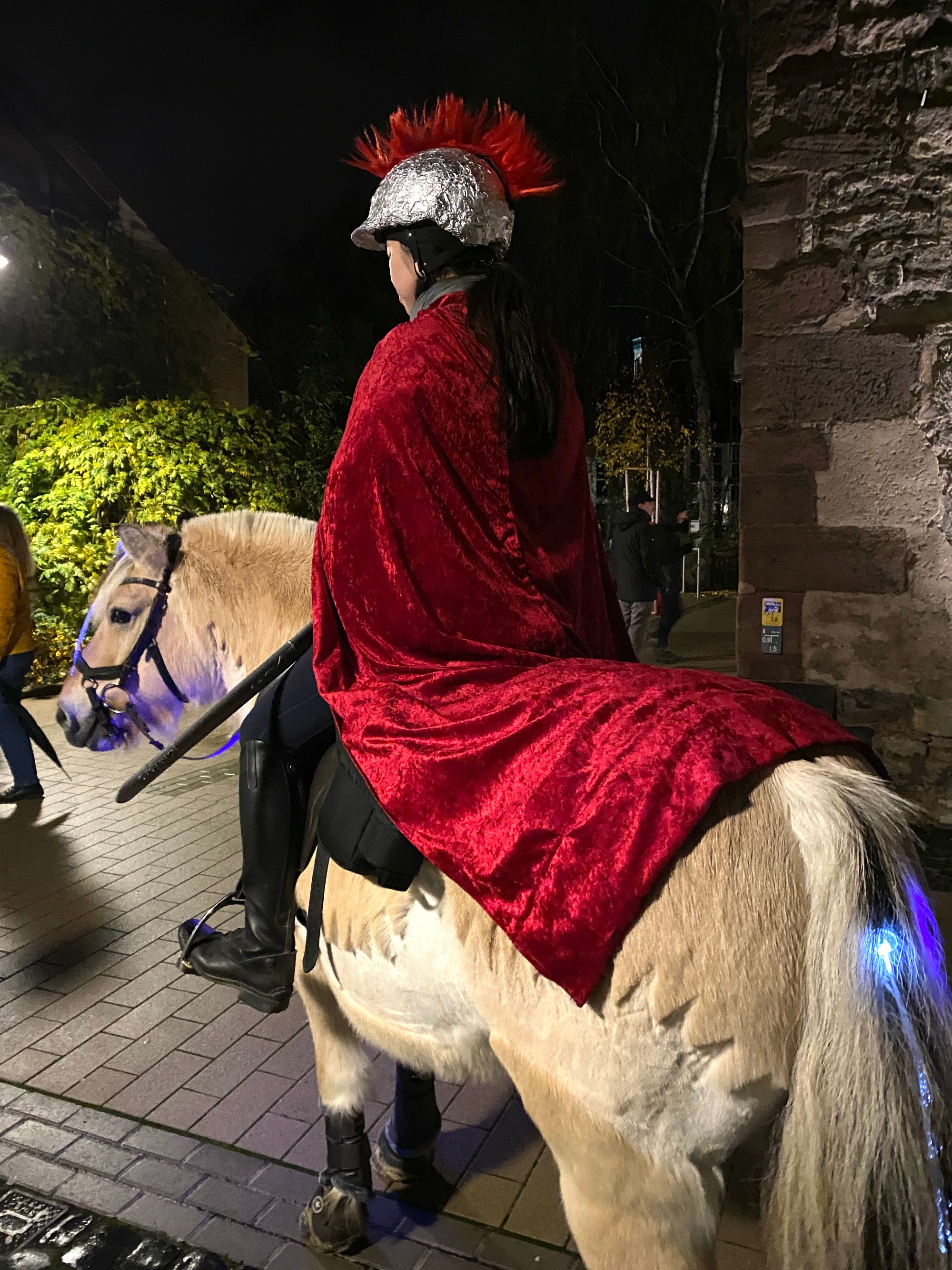 Person im roten Umhang und silbernem Helm reitet bei Nacht auf Pony durch eine Straße.